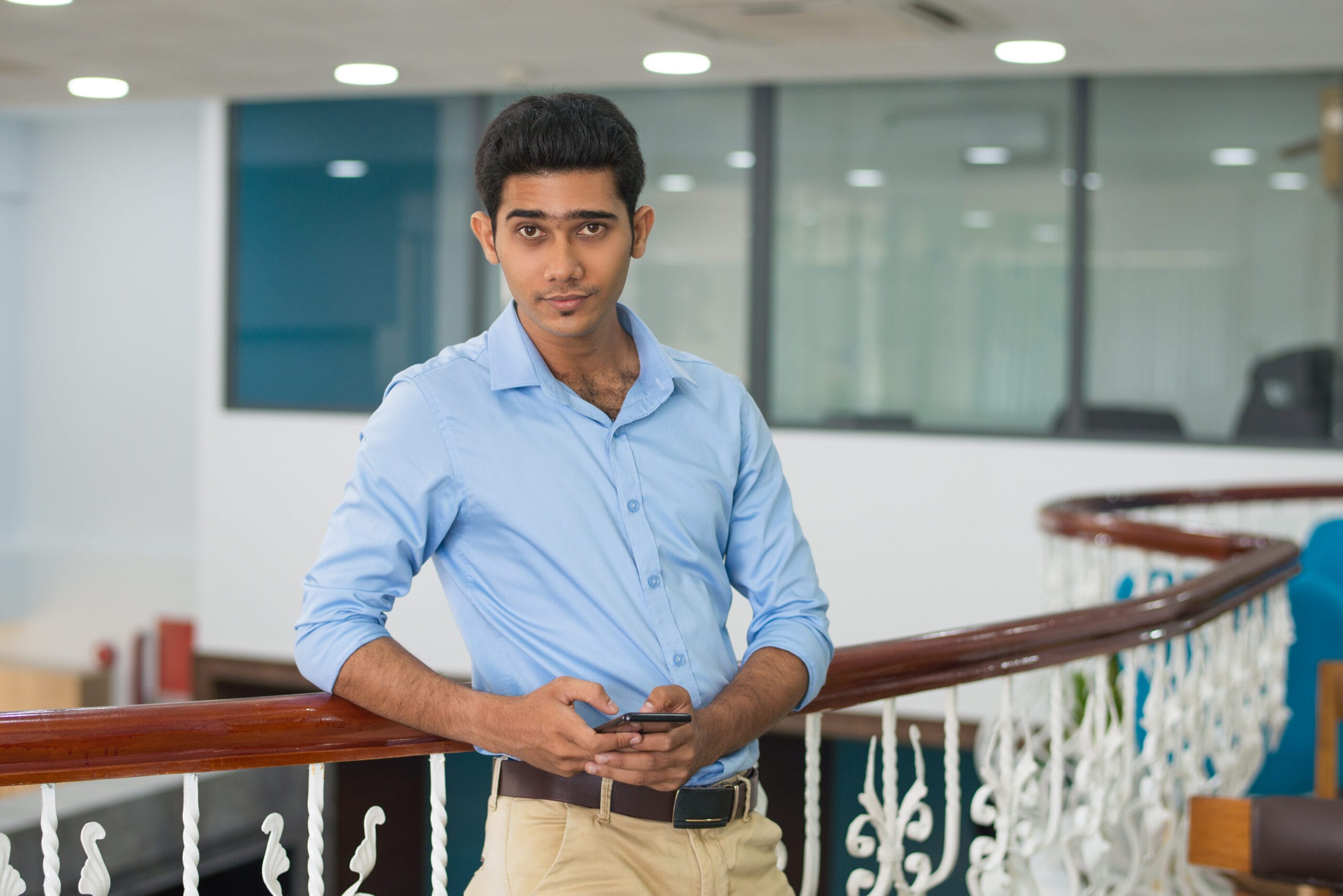 Content confident young man leaning on railing in modern office. Handsome Indian businessman in blue shirt using smartphone and looking at camera. Entrepreneur concept